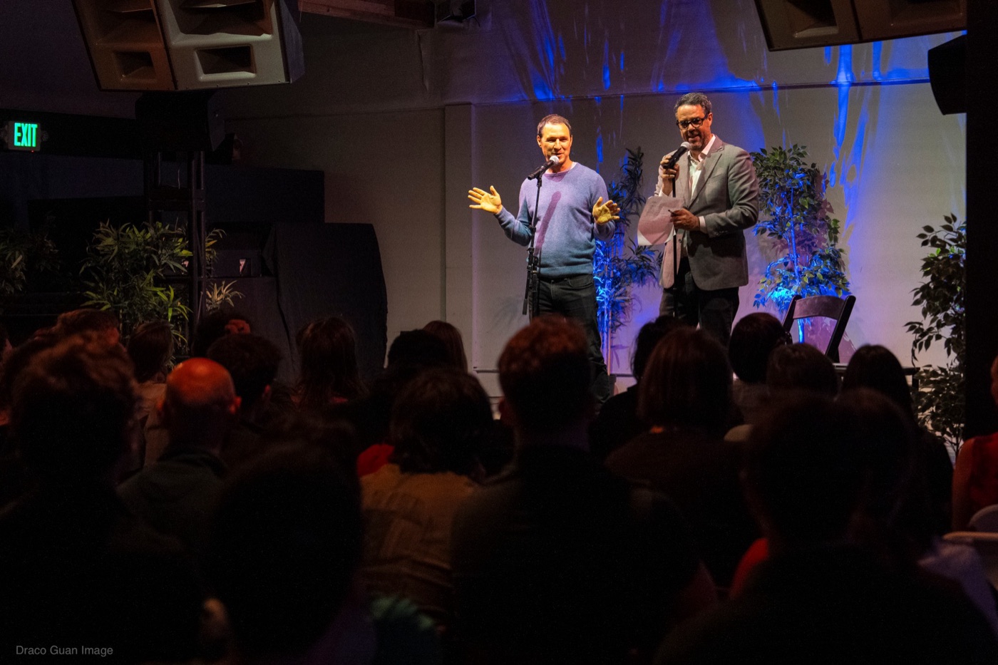 Misha on stage with a microphone beside a guest speaker, in front of a packed audience.